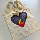 Ein Werk: helle Stofftasche verziert mit schwarzer Brailleschrift und gezeichnetem Flammenherz in Schwarz-Rot-Gelb.