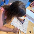 Eine Frau verziert mit dem Dot-Pen eine Stoff-Tasche. Auf der Tasche ist schon ganz viel in verschieden farbiger Brailleschrift etwas geschrieben.