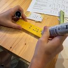 Kinderhände machen mit einem Dot-Pen dicke Punkte auf einen Filz-Schlüsselanhänger. Dadurch entsteht vergrösserte Brailleschrift.