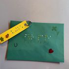 Zwei Werke: Ein gelber Schlüsselanhänger mit dunkelgrüner Brailleschrift verziert. Daneben liegt ein dunkelgrünes Kuvert, welches mit Brailleschrift verziert ist.